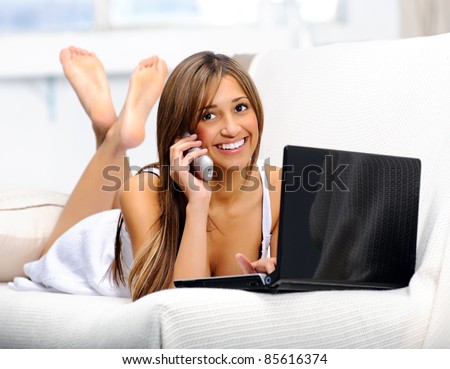 Woman lying on the couch using her laptop and having a happy conversation on the telephone