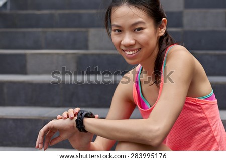 portrait of sporty asian chinese woman sitting outdoors checking workout time on wearable technology watch after run