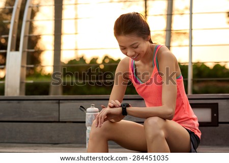 sporty asian chinese woman sitting outdoors checking workout time on wearable technology watch after run