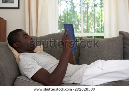 african black man relaxing on sofa couch reading literature novel story book at home living room lounge
