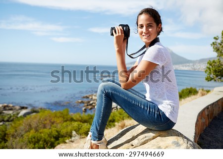 young, attractive woman sitting on a rock, poses for a pic near the sea with digital camera in her hand