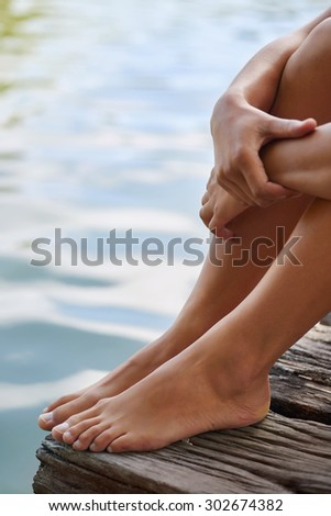 Close up of female feet as she relaxes by the lake sitting on the edge of a wooden jetty, hands over her knees