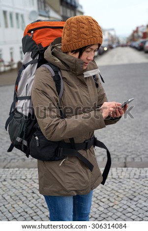 Young Asian backpacker tourist woman in rain jacket and beanie busy with her mobile Phone middle of urban city street