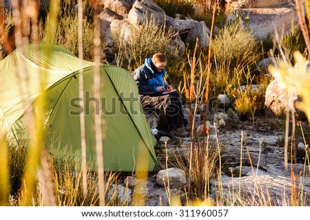 Adventure camping man cooking alone outdoors with tent, sunrise and lens flare in the mountain morning sunlight. Happy explorer enjoying a meal in the wilderness