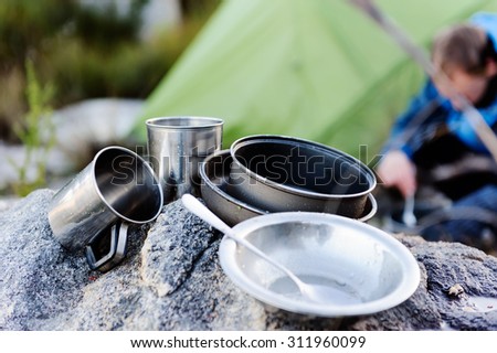 Camping cooking gear, pots and utensils in focus while an adventure man cooks food in the background next to an extreme weather tent outdoors in the mountains.
