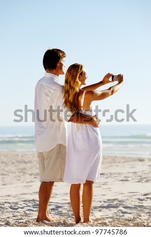 Summer beach honeymoon couple standing on the sand and taking pictures of the beautiful sunset over the ocean