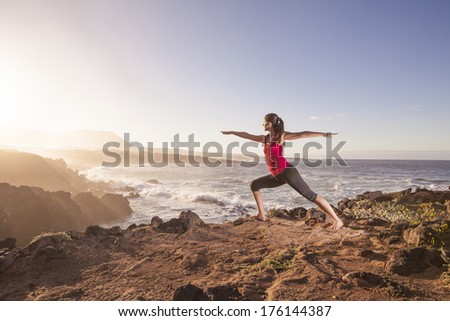 Young woman practicing yoga warrior pose near the ocean at sunset