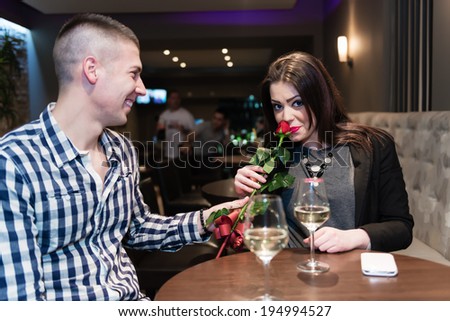 Young adults in cafe celebrating. Man give rose to his love. She smelling rose and look at camera.