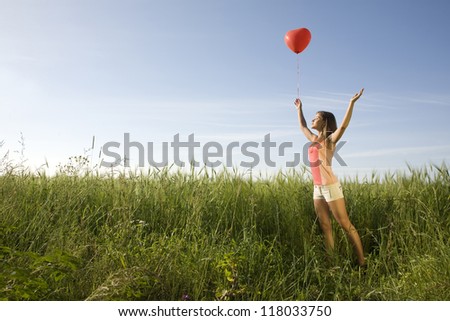 Young woman standing on the field with the red heart-shaped balloon and her arms raised