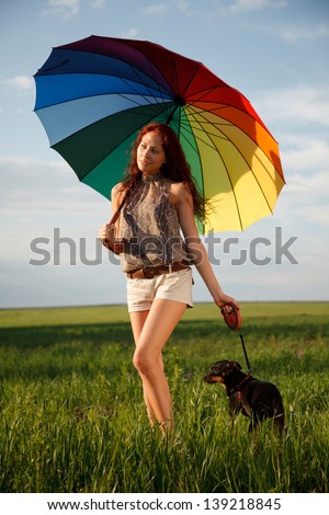 Young slim woman is walking on the field with the dog under big colorful umbrella
