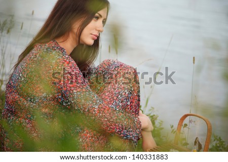 Beautiful girl in colorful dress sits near water. Grass out of focus on the foreground