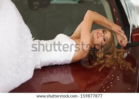 Young sensual bride lies on the red car bonnet and shows emotions