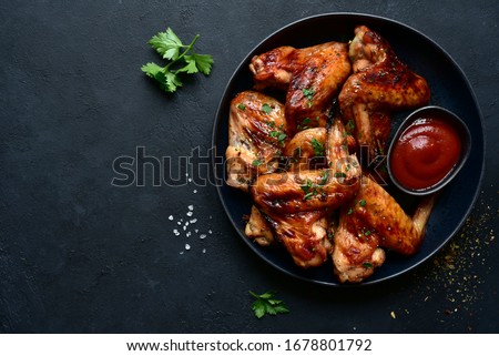 Grilled spicy chicken wings with ketchup on a black plate on a dark slate, stone or concrete background. Top view with copy space.