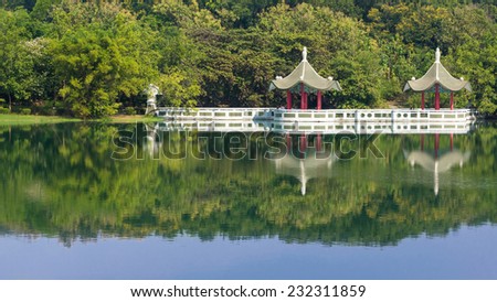 A Scene of Cheng Ching Lake With Two Chinese Pavilions by The Lake. The Water Reflect Green Forest , The Pavilions And Blue Sky. With Tourists Walking by The Lake