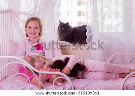 Cute funny little girls (sisters) play in bed, on the window sits a cat. Selective focus.