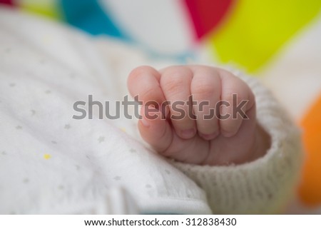 Close-up of the closed left hand of a baby who wears a fleece jacket, in front of a colorful background.