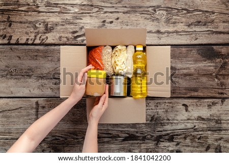 Person woman receiving donation Food box. Female volunteer hands packing donation box with food items of staple products on wooden table. Donate Food delivery concept. Donations grocery canned food. 商業照片 © 