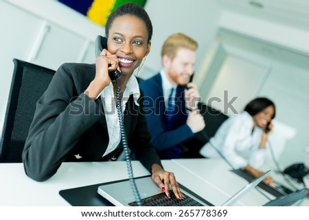 A beautiful, black, young woman working at a call center in an office with her red haired partner on the other end of the desk talking to another customer