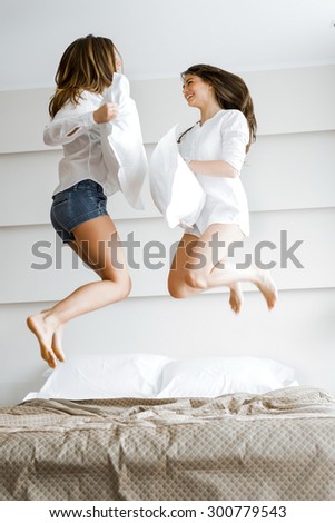 Two beautiful women jumping from joy on bed and fighting with pillows in a luxurious hotel room