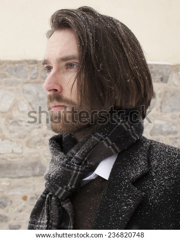 Side profile of a bearded adult male with long hair against a stone wall during a blizzard.