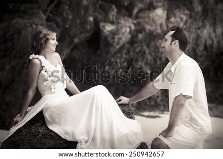handsome groom looks at beautiful bride sitting on the rock at the sand beach