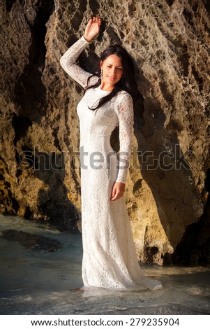 brunette bride in white wedding dress leans on large cliff in water