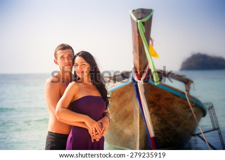 brunette young girl and handsome guy hug against longtail boat at sunrise