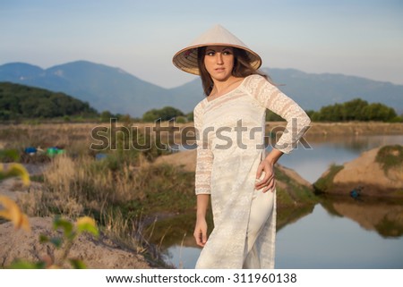 slim blonde girl in Vietnamese national white long dress and hat smiles against lakes country house and mountains