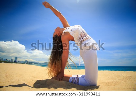 blonde girl in white lace costume in yoga asana wheel back bend on beach against blue sky