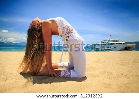blonde girl in white lace in yoga asana back bend camel on beach against blue sky and tourist vessel