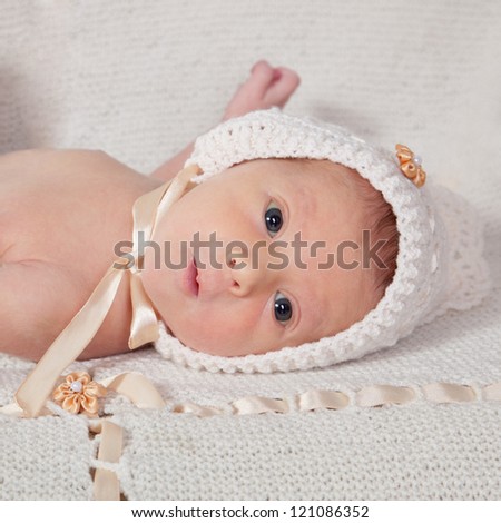 10 day old newborn baby in white fashion knitting hat laying on the tummy on white knitting blanket looking very curious. Selective focus on baby face