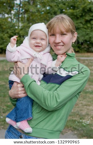 Young happy mother dressed in green polo-neck sweater holding her eight month old baby laughing daughter dressed in pink hooded cardigan and jeans outdoors