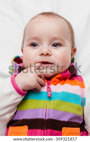 Cute baby in striped colorful autumn  spring outfit sucking finger while lying on the back on white sheet background. Portrait, closeup.