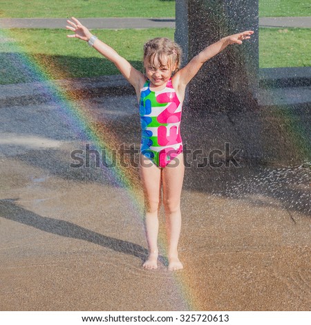 Smiling little girl standing in streams of city splash pad fountains. Kid happy to be wet and fresh. Rainbow in the fountain.