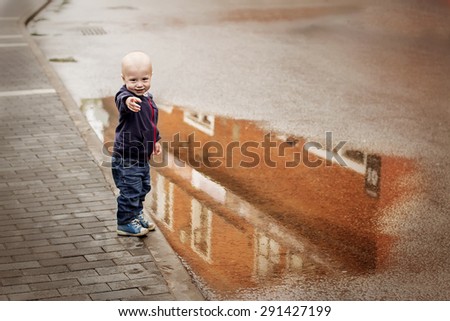 Little boy pointing his finger on you standing near the big puddle on the ground with reflection of red building. Image with selective focus