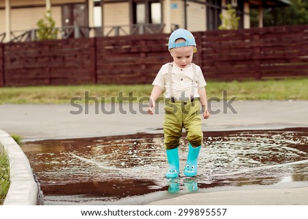 Cute little boy in cap and blue rubber boots is having fun splashing through the puddles. Image with selective focus