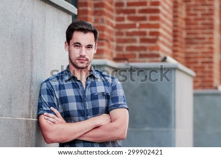 Portrait of attractive dark-haired young man standing crossing arms against an urban background. Image with selective focus