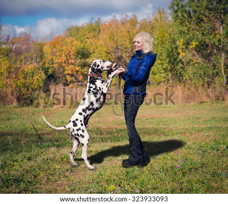 Beautiful blonde girl in a blue jacket and black jeans dances with her dog breed Dalmatian in nature among green grass and trees, they are happy and smiling