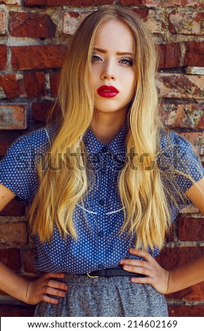 Emotive portrait of a young beautiful blond-haired girl wearing trendy blue shirt and grey skirt, posing over old brick wall.  Healthy skin with freckles, glossy hair. Outdoor shot