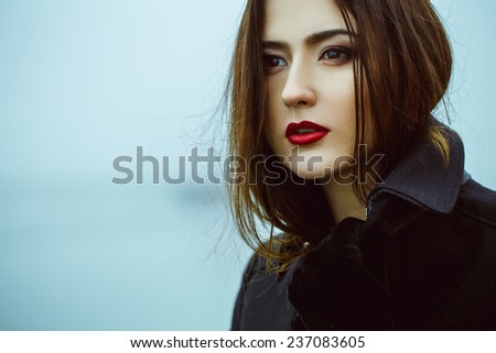 High fashion concept. Emotive portrait of beautiful brunette with long curly hair and perfect make up wearing black coat. Windy and misty weather. Italian luxurious style. Outdoor shot