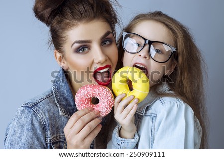 Two beautiful sisters (girls)  in hipster style clothes and in glasses eat cupcakes  at studio.  Perfect skin and make up.  Casual clothes.