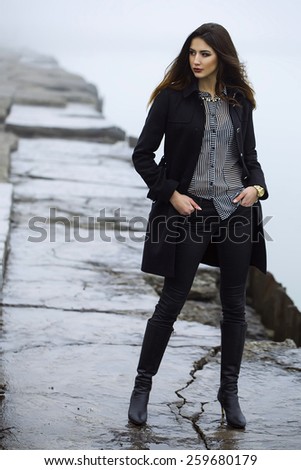 High fashion concept. Emotive portrait of beautiful brunette with long curly hair and perfect make up wearing black coat. Windy and misty weather. Italian luxurious style. Outdoor shot