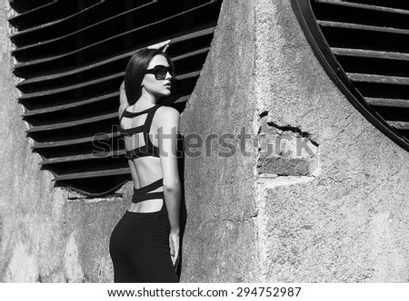 Fashion portrait of young beautiful brunette girl in sexy black dress and sunglasses. Outdoor shot. Black and white photo. Urban style