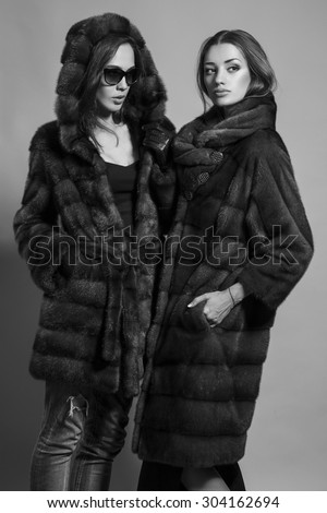 Fashion shot of two elegant beautiful girls (brunette and blonde) in studio on grey background wearing sunglasses and furs coat . Shopping inspiration. Monochrome (black and white)  photo