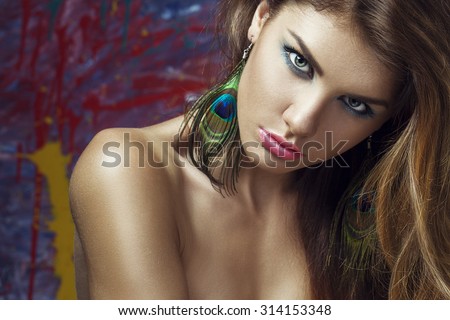 Sexy Beauty Girl with pink Lips, Green Eyes and earrings made of peacock feathers. Provocative blue Make up. Luxury Woman posing on art background, the girl is the artist\'s Muse. Gorgeous Woman Face.