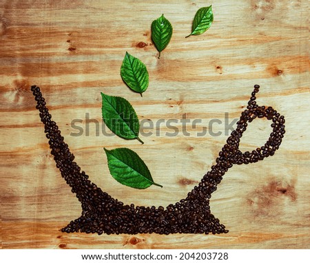 Coffee beans cup concept. Imbue beans of black coffee make a form of a cup with fresh green leaves over wooden texture background. Studio shot. Copy space.