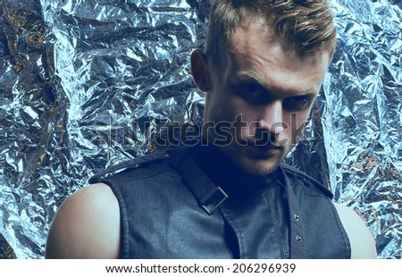 Arty portrait of young handsome blue-eyed man with trendy haircut with strong intense look in sleeveless zip-front vest of dark jeans posing over silver foil background. Close-up. Studio shot