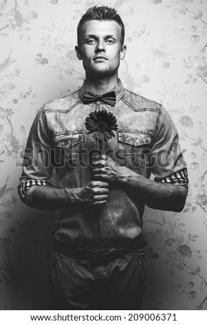 Arty black and white fashion portrait of young and handsome hipster man in jeans shirt, pants and bow-tie with a flower over vintage background. Pensive emotive face. Close up. Studio shot