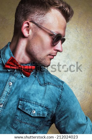 Eyewear concept. Handsome male model with in trendy glasses, jeans shirt and red bow-tie over golden background. Clean healthy skin. Hipster & vintage (retro) style. Close up. Studio portrait