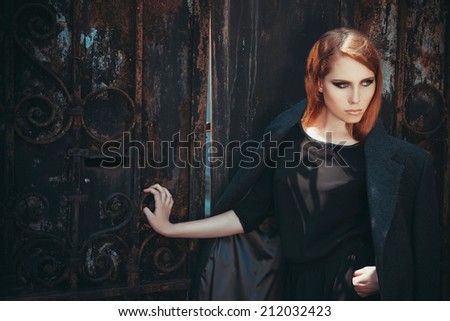 Vogue fashion concept. Portrait of young beautiful red-haired girl in trendy black dress and grey coat posing over old gothic gate. Perfect skin and strong smoky eyes make-up. High fashion style.